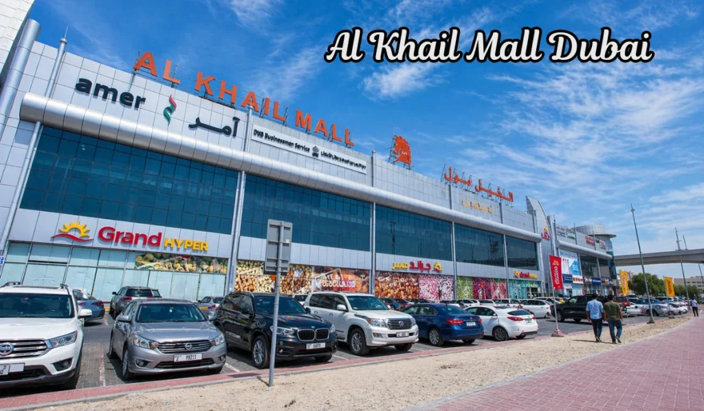 Wide daytime view of Al Khail Mall with parked cars, bright blue sky, storefront signs, and two people walking along the sidewalk.