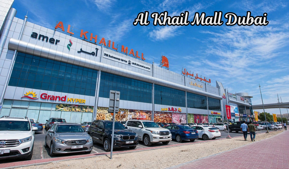 Wide daytime view of Al Khail Mall with parked cars, bright blue sky, storefront signs, and two people walking along the sidewalk.