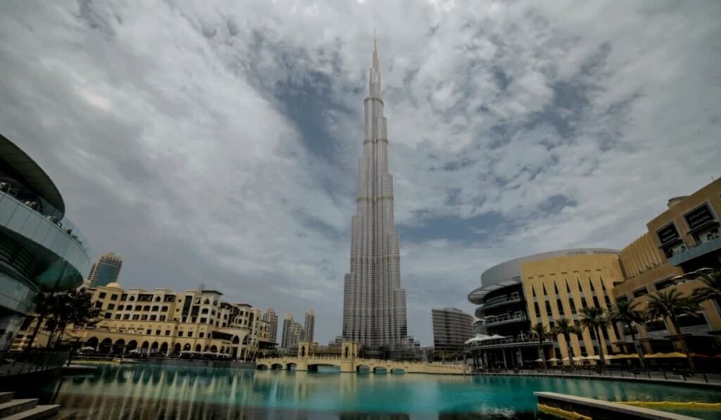 Burj Khalifa rises over a turquoise reflecting pool in Dubai, framed by modern buildings beneath a dramatic cloudy sky.