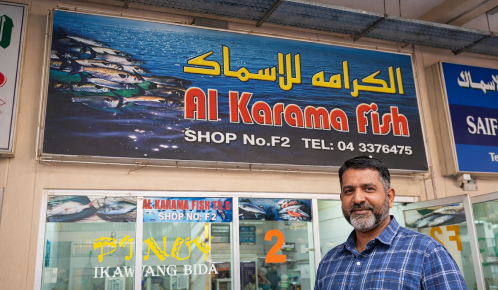 A man standing outside the Al Karama Fish shop in Dubai, featuring a large bilingual sign and storefront windows.
