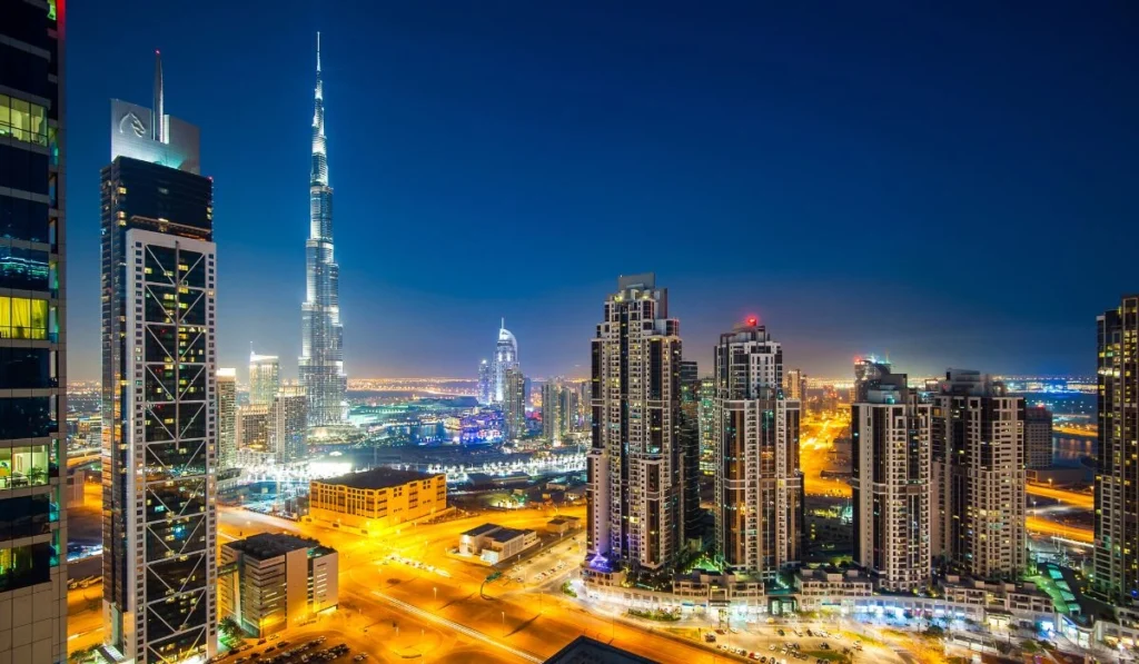 Night skyline of downtown Dubai with the illuminated Burj Khalifa rising above surrounding high-rise towers and glowing roads.