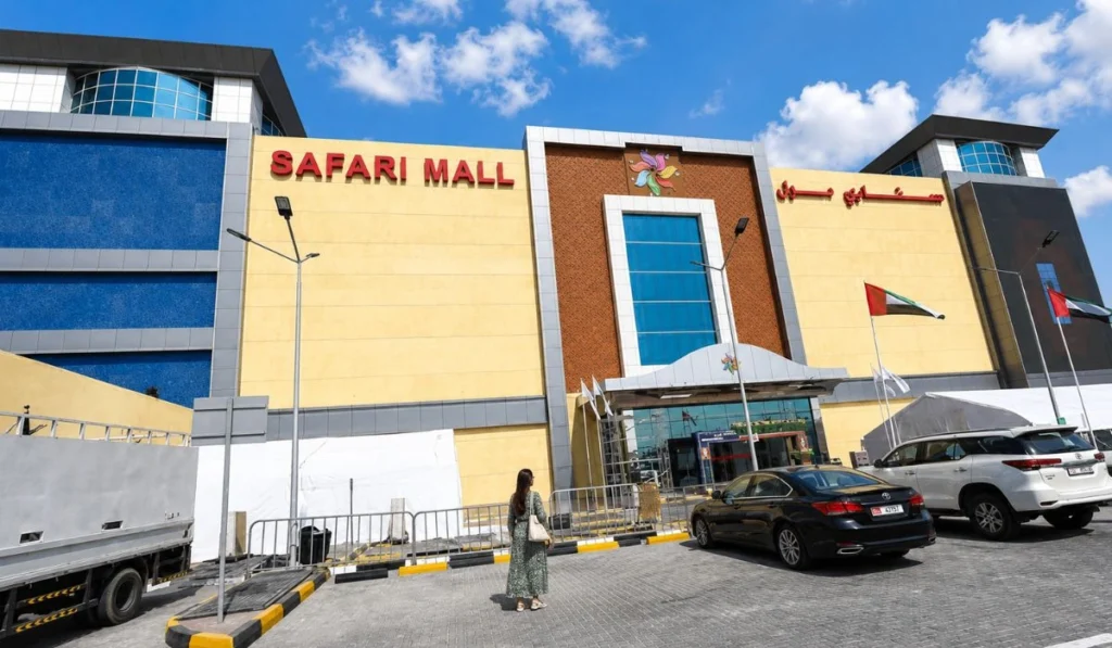 Safari Mall Sharjah entrance with clear blue sky, clouds, parking area, black car and woman standing outside the mall.